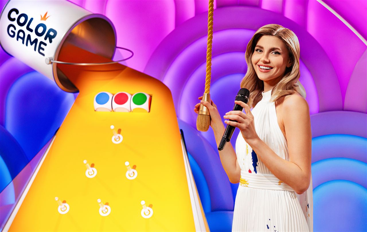 Color Game Bonanza presenter in a white dress holding the release cord beside the pin-lined yellow ramp and three colour dice