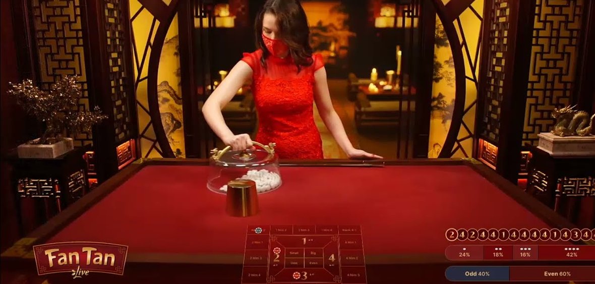 A fan tan live table with the glass dome over the bead pile and the metal cup, with the dealer and the Asian themed studio behind