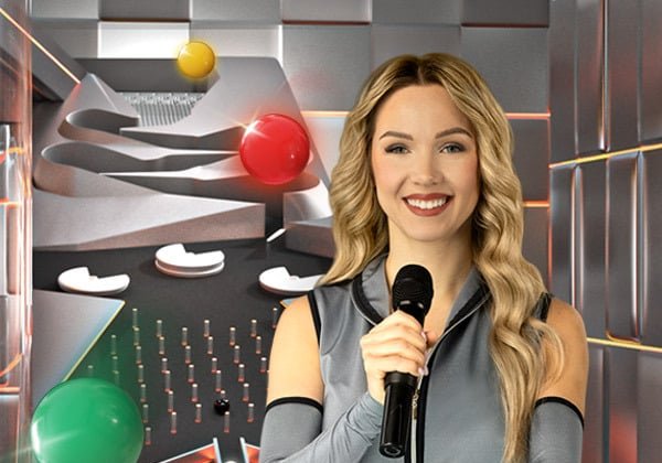 Marble Race Live host with a microphone beside the obstacle track as red, yellow and green marbles roll down