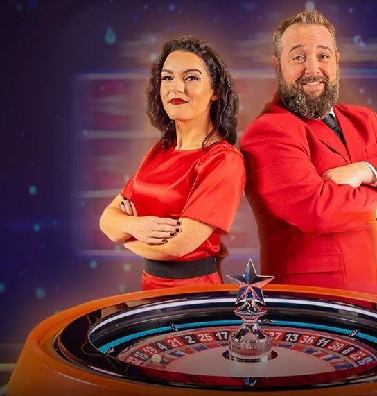 Two Mega Fire Blaze Roulette presenters in red behind the wheel with its star-topped turret