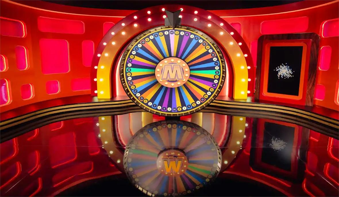 The mega wheel live studio wheel with 54 numbered segments from 1 to 40, the M hub and the lit arch reflected in the glossy floor