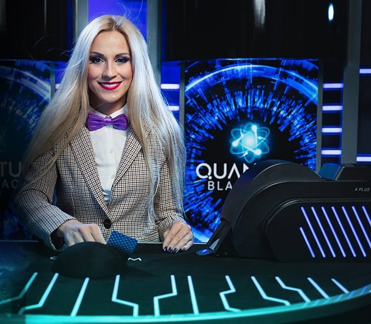 Quantum Blackjack dealer in a purple bow tie at the Quantum table, with the Quantum Blackjack screen and card shuffler behind